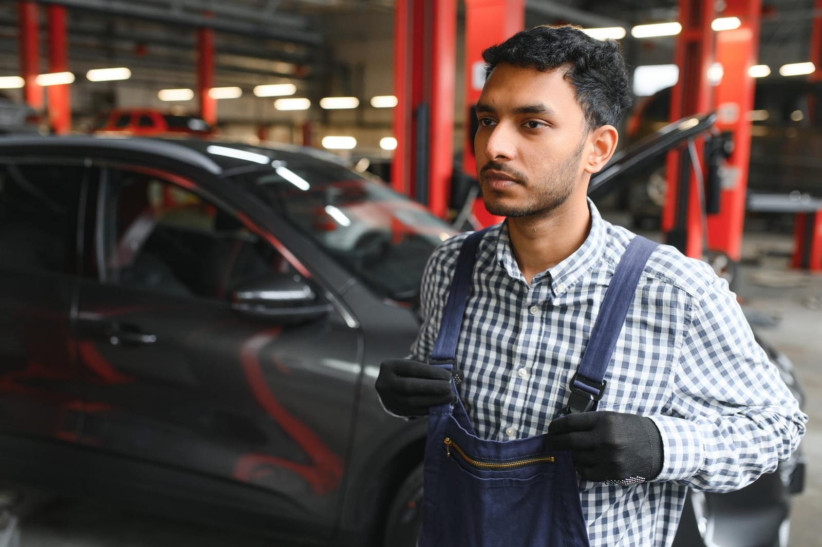 Mecânico de macacão ao lado de um SUV preto dentro de uma oficina com elevadores vermelhos.