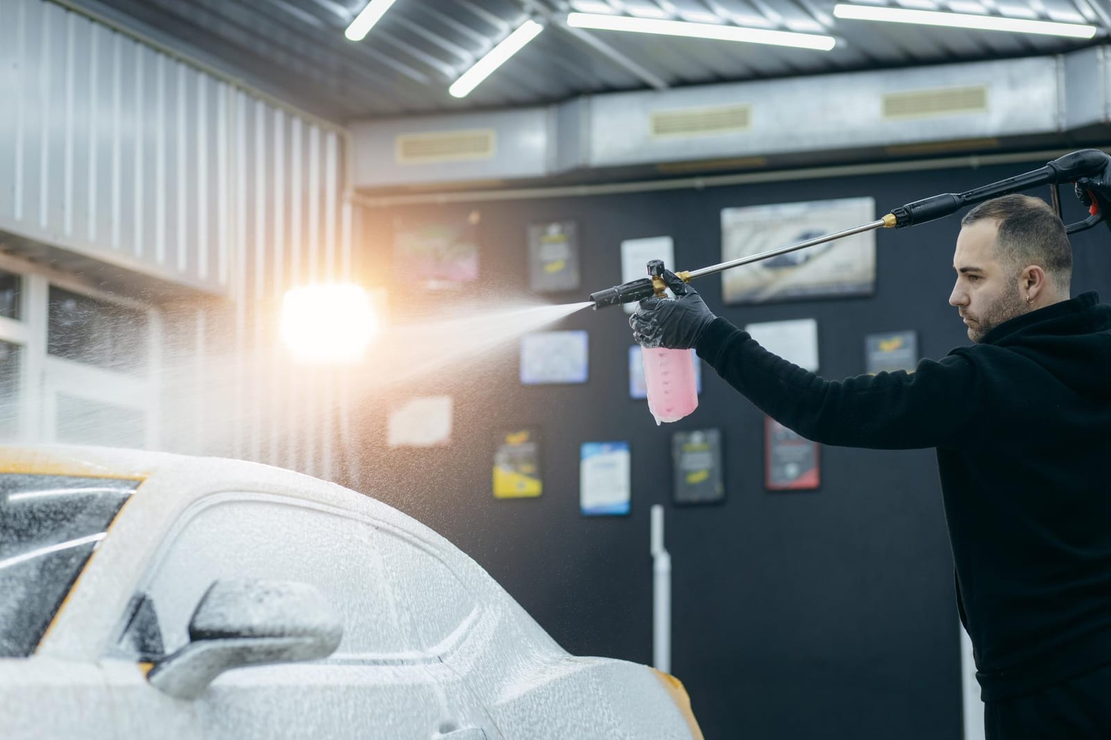 Profissional de estética automotiva aplicando espuma de limpeza sobre um carro em um box de detalhamento.